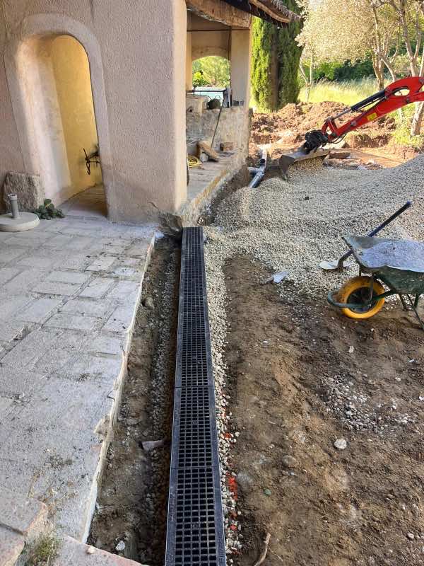 Caniveau à grille posé au bord d'une terrasse dallée, gravier et mini-pelle en arrière-plan
